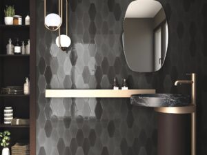 Bathroom with dark gray variegated elongated hexagon tile wall; gold shelf and faucet, stone patterned sink basin, mirror, light fixtures, and floor to ceiling shelving