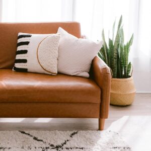 Tan leather couch with white decorative pillows; white backlit curtains; snake plant in a basket; pale floors; white and black shag rug