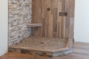 Shower partially installed; pebbled tile floor, wood-look tile up one wall, ledger stone at right angle; wood-look flooring outside of shower