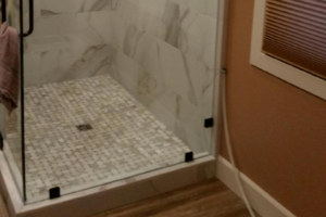 Corner of a shower showing basketweave mosaic tile floor and stone-look walls; outside of shower is woodlook tile flooring