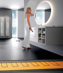 woman standing in a bathroom with dark tile floor, at front edge the underlayment is exposed showing orange membrane and inlaid cables