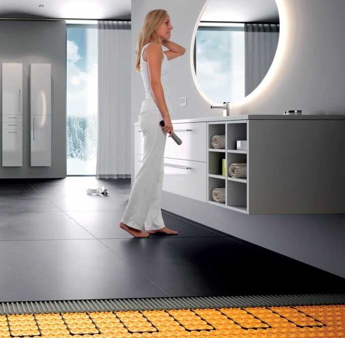 woman standing in a bathroom with dark tile floor, at front edge the underlayment is exposed showing orange membrane and inlaid cables